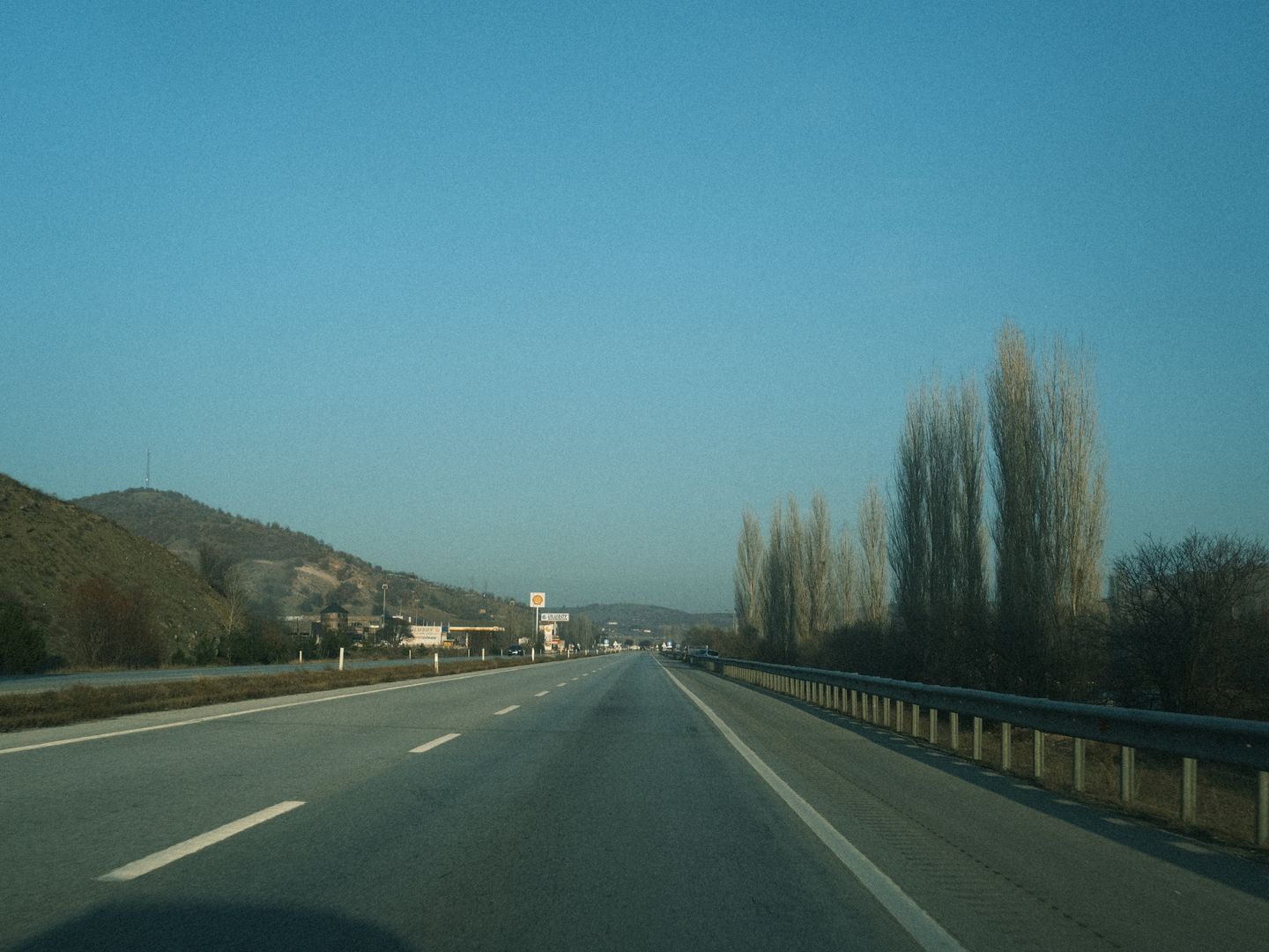 Expansive view of a highway stretching into the distance, flanked by sparse trees and hills under a clear blue sky.